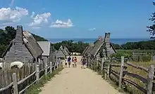 A modern-day photograph of a village consisting of small, primitive wooden houses.  Most of the houses have thatched roofs.  In the distance is a large expanse of ocean and a clear blue sky.