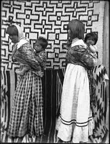 A black and white photograph of two Native American women with children in backpacks