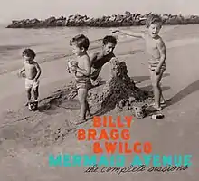 A black-and-white photo of a man building a sand castle on a beach with several children standing around him, one of whom is pointing toward the body of water. Superimposed text reads "BILLY / BRAGG / & WILCO" in orange, "MERMAID AVENUE" in blue, and "the complete sessions" in black script.