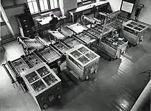 Looking down from above at a room at an intricate mechanical device which fills the room. In the background, a man sits at a desk next to a filing cabinet. Owens 1991, p. 15.