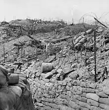 A black-and-white photograph of damaged field defences in the daylight: A deep sandbag-lined trench runs from the bottom right to the bottom left away from the camera, while further up the slope to the right, disturbed earth, damaged star pickets, and wire entanglements and other debris are evident below the ridgeline.