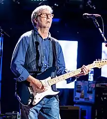 A man on a stage wearing a blue shirt. He is holding a blue guitar behind a microphone.