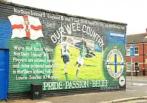 A large mural on a wall. Prominent features include the Ulster Banner, the IFA logo, the scoreline "Northern Ireland 1, England 0", and the captions "OUR WEE COUNTRY" and "PRIDE – PASSION – BELIEF"