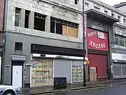 Grey stone-fronted building with blackened empty windows and shutters down. Graffiti reads "Who burnt us out? People before profit" - and "Fat cats get fatter, puts the kittens out in the cold."