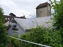 Very close view of a low iron church with a corrugated iron roof and a stone tower.  This a low brown cap-style roof and a projecting crucifix..